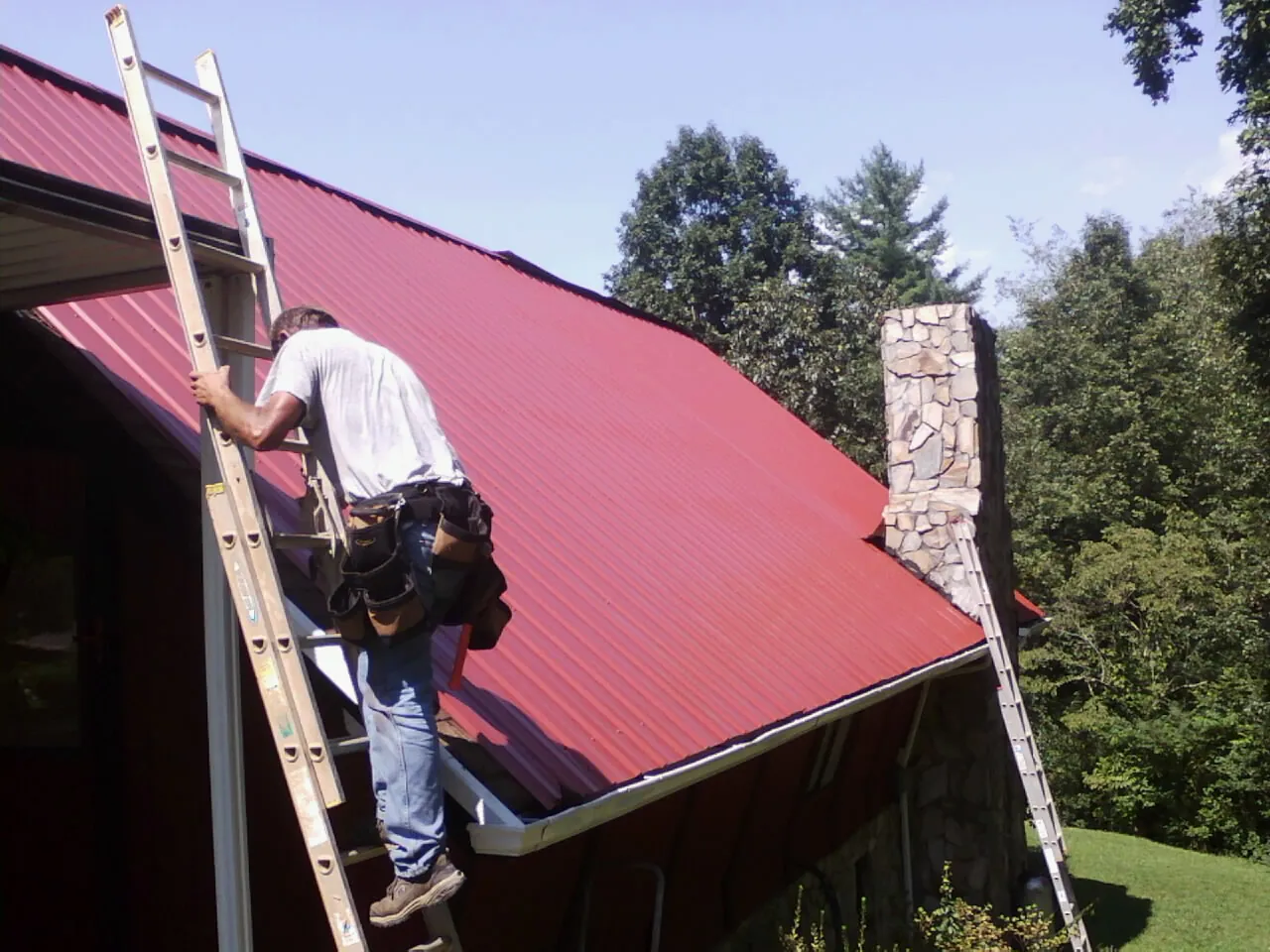 Roofer on a ladder beside a red metal roof and stone chimney — Soffit & Fascia Repair by Roofing Brothers in Kenyon